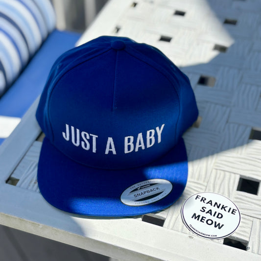 Blue cap with 'JUST A BABY' text on a white surface by Frankie Said Meow